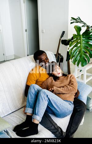 Cheerful multiracial couple in casual clothes relaxing on sofa while spending time together in living room at home Stock Photo