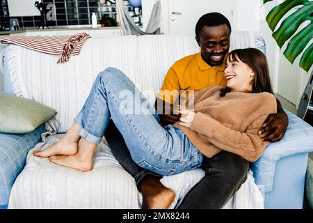 Cheerful multiracial couple in casual clothes relaxing on sofa while spending time together in living room at home Stock Photo