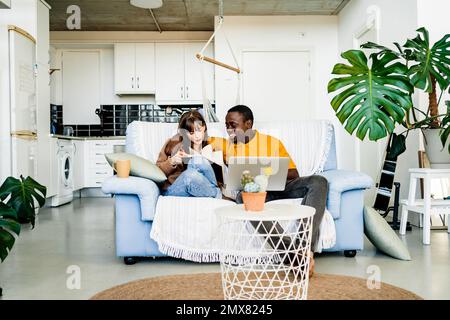 Happy multiethnic couple in casual clothes sitting on sofa reading book and using laptop in living room at home Stock Photo