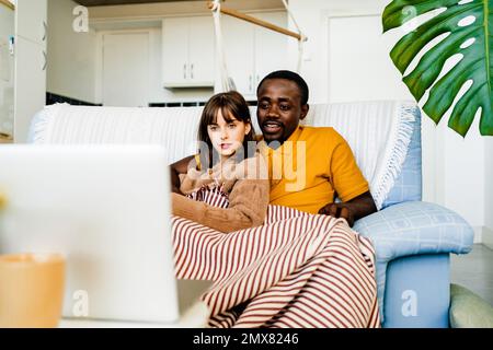 Young multiracial couple in casual clothes sitting on sofa and browsing laptop while spending leisure time at home Stock Photo