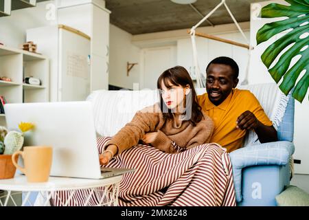 Young multiracial couple in casual clothes sitting on sofa and browsing laptop while spending leisure time at home Stock Photo