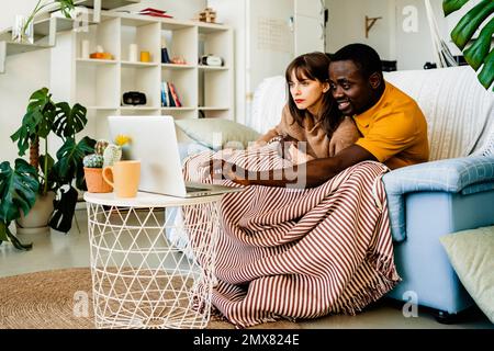 Young multiracial couple in casual clothes sitting on sofa and browsing laptop while spending leisure time at home Stock Photo