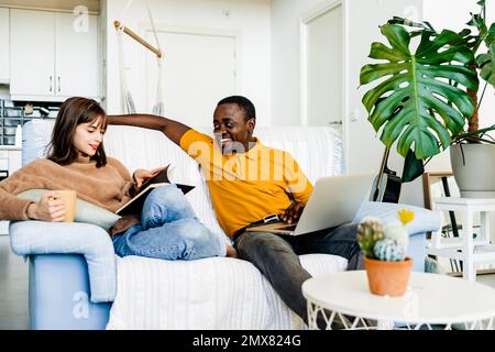 Happy multiethnic couple in casual clothes sitting on sofa reading book and using laptop in living room at home Stock Photo