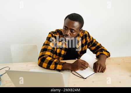 Focused pensive African American male freelancer in casual clothes writing notes in notebook while working on remote project on laptop Stock Photo