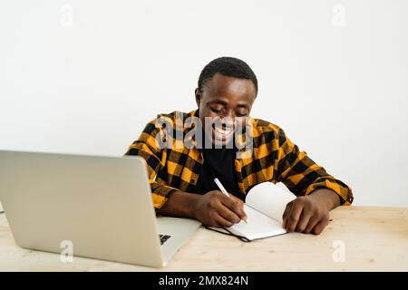 Positive African American male freelancer in casual clothes writing notes in notebook while working on remote project on laptop Stock Photo