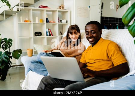 Happy multiethnic couple in casual clothes sitting on sofa reading book and using laptop in living room at home Stock Photo