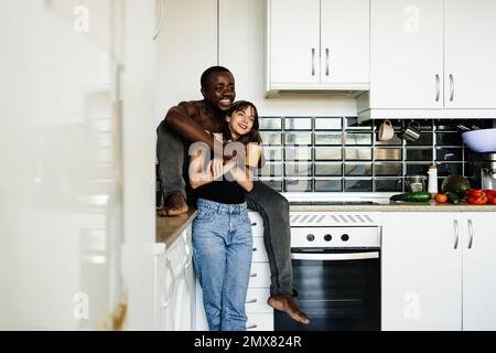 Cheerful diverse couple in casual clothes hugging and smiling looking awayin modern kitchen at home Stock Photo