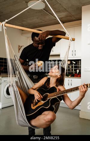 Positive young female musician playing acoustic guitar while sitting on swing chair and looking at black boyfriend with cup of beverage during weekend Stock Photo