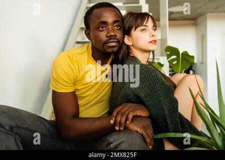 Thoughtful young multiethnic couple in casual clothes and embracing while sitting together on staircase with green plant at home Stock Photo