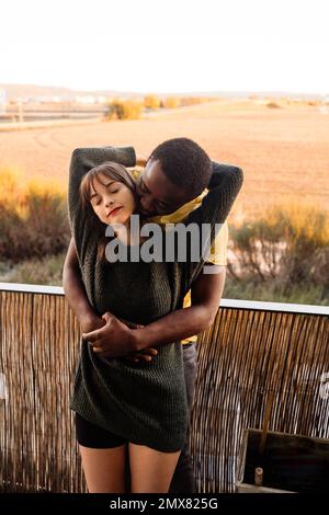 Delighted young multiracial couple in casual wear cuddling while standing with eyes closed on terrace of house and enjoying weekend in countryside Stock Photo
