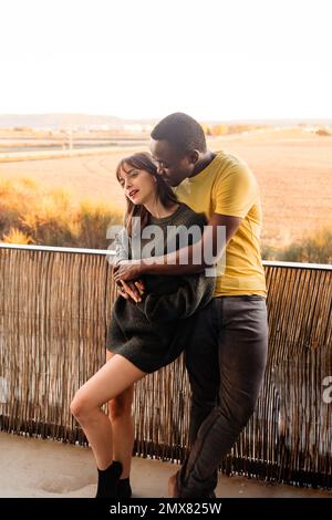 Delighted young multiracial couple in casual wear smiling and hugging while standing on balcony of house and leaning on railing against sunset sky Stock Photo