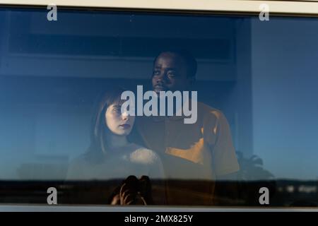 Through glass of calm thoughtful multiracial couple in casual wear standing looking away while spending time at home near window Stock Photo