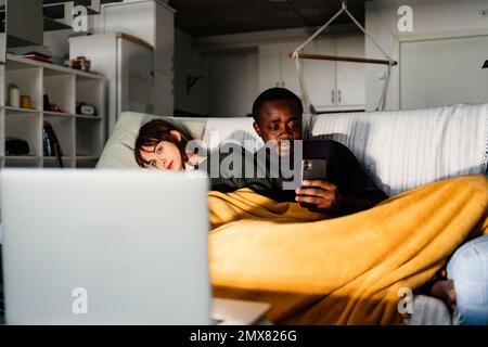 Concentrated African American male checking social media while resting on sofa under blanket with girlfriend watching movie on laptop in living room Stock Photo