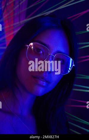 Portrait of serious young female in trendy glasses looking at camera while standing in neon lights Stock Photo