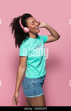 side view of happy young African American female in casual clothes with headphones while standing against pink background Stock Photo