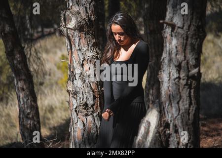 Thoughtful young Hispanic female tourist with long dark hair in black casual outfit and looking down while standing amidst trees on sunny day in fores Stock Photo