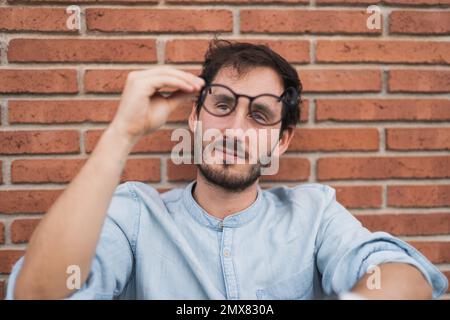 Pensive bearded man in casual shirt sitting near brick wall with eyeglasses and looking away Stock Photo