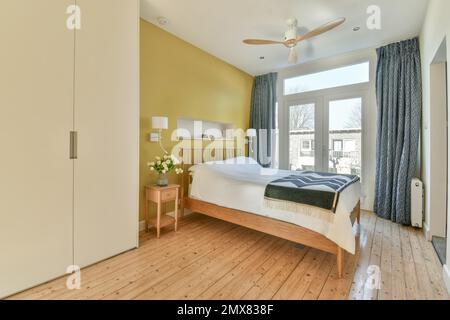 Interior design of contemporary bedroom with wooden floor and bed against yellow wall near big window with blue curtains bedside table with flowers an Stock Photo