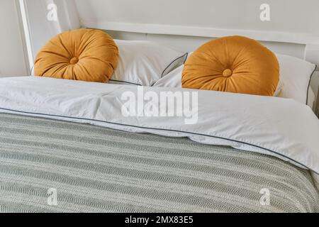 Soft bed with pillows and blanket decorated by orange cushions placed near white wall in light bedroom at home Stock Photo