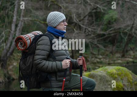 Pensive senior male hiker in warm clothes and hat with backpack and trekking poles walking on pathway in forest during hiking trip Stock Photo