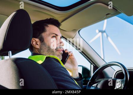 Side view low angle of bearded man in light green vest sitting on driver seat against modern wind turbine and talking via mobile phone Stock Photo