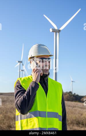 Smiling bearded male worker in vest and safety helmet standing on countryside roadway against windmills while speaking via cellphone Stock Photo