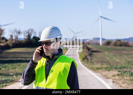Serious bearded male worker in vest and safety helmet standing on countryside roadway against windmills while speaking via cellphone Stock Photo