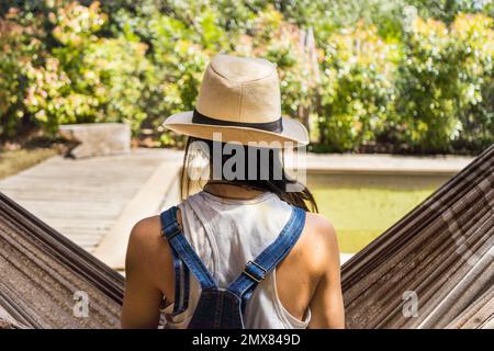 Back view of anonymous female in hat and denim jumpsuit sitting on hammock near swimming pool and trees while resting during weekend Stock Photo