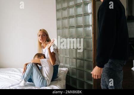 Portrait of scared mother and daughter hugging each other sitting on sofa while aggressive unrecognizable father angrily yelling at them at home. Stock Photo