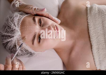 A young woman receives an injection of anti-aging botox filler in the forehead by a professional beautician in a beauty salon, rear view. Facial treat Stock Photo