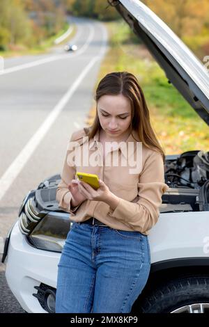 A woman waits for assistance near her car broken down on the road side. Woman with a broken car with open hood. Upset young woman with cell phone near Stock Photo