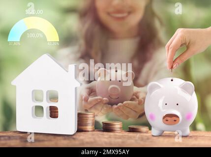 Woman putting coins into piggy bank at table, closeup. Collecting money to buy house Stock Photo