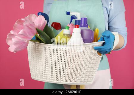 Woman holding basket with spring flowers and cleaning supplies on pink background, closeup Stock Photo