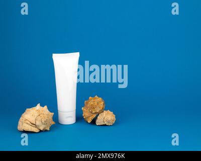 White tube and whelk seashells on blue background, copy space. Mockup plastic tube for sunscreen, nourishing or moisturizing cream Stock Photo