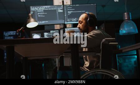 Young engineer with chronic disability working on IT software coding, using source code and data. Male coder wheelchair user with impairment developing system interface. Handheld shot. Stock Photo