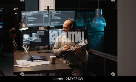 App developer playing air guitar in coding office, enjoying music and song late at night. Having fun fooling around, feeling cheerful and careless while he programs new code. Handheld shot. Stock Photo