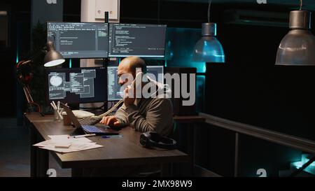 Male app developer answering landline phone call late at night, talking on office telephone with cord and typing source code. Creating algorithm for agency, writing html script. Handheld shot. Stock Photo