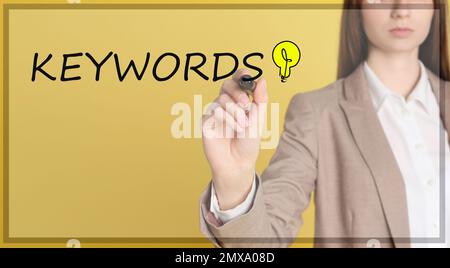 Woman writing word KEYWORDS on transparent board against yellow background, closeup Stock Photo