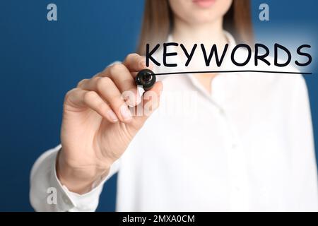 Woman writing word KEYWORDS on transparent board against blue background, closeup Stock Photo