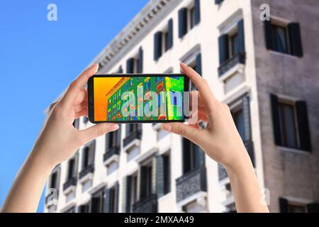 Woman detecting heat loss in building using thermal viewer on smartphone, outdoors. Energy efficiency Stock Photo