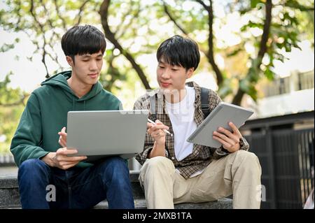 Two smart young Asian male college students focusing on his school project, looking at laptop screen, using laptop and tablet, discussing and working Stock Photo