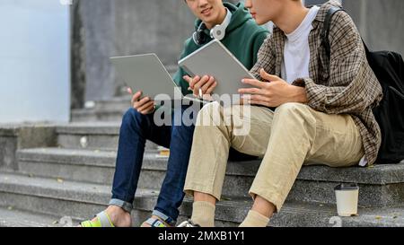 Cropped shot of two Asian male college students in casual clothes sitting on street stairs with his portable tablet and laptop, talking and spending t Stock Photo