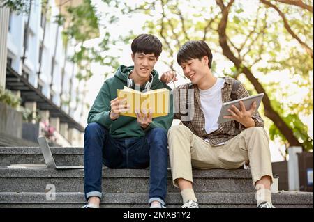 Two young Asian male college students relax outside of the campus building, reading a book together, preparation for an exam. Stock Photo