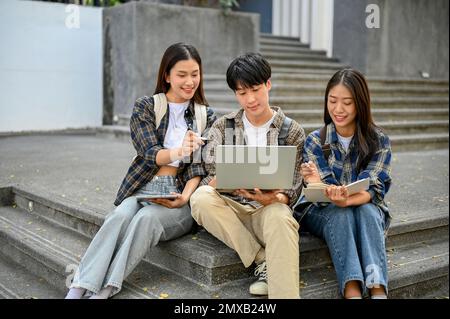 Group of young Asian college students sitting on stairs in front of the campus building, talking and focusing on their school project, sharing ideas, Stock Photo