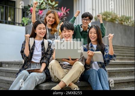 Group of cheerful young Asian college students sitting on stairs in front of the campus building, showing fists, clenched fists, celebrating triumph, Stock Photo