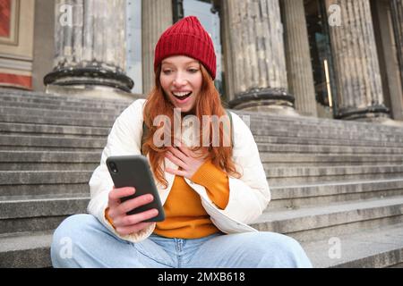 Redhead female tourist talks on mobile phone, video chatting with friends, says hi and waves hand at smartphone camera. Stock Photo