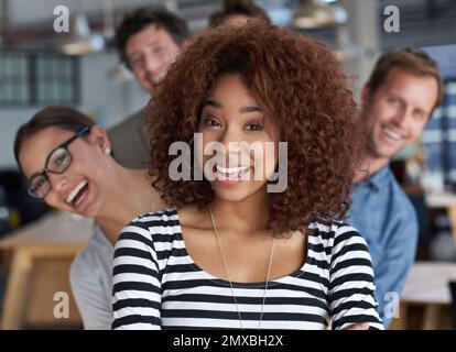 We have a multifaceted approach to business. Closeup portrait of a smiling young woman with friendly coworkers peeping out behind her. Stock Photo