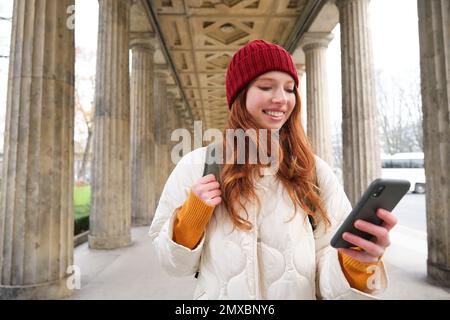 Mobile broadband and people. Smiling redhead 20s girl with backpack, uses smartphone on street, holds mobile phone and looks at application. Stock Photo