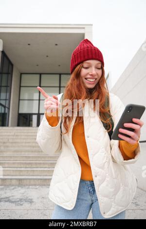 Vertical shot of redhead girl tourist, shows her friend via video chat building, points somewhere, talks on mobile phone. Stock Photo
