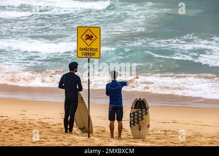 Two male surfers stood beside dangerous current sign on a Sydney beach Australia holding surfboard and bodyboard Stock Photo
